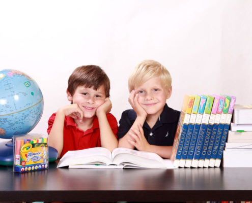 two kids sit at a table with books, crayons, and other school supplies